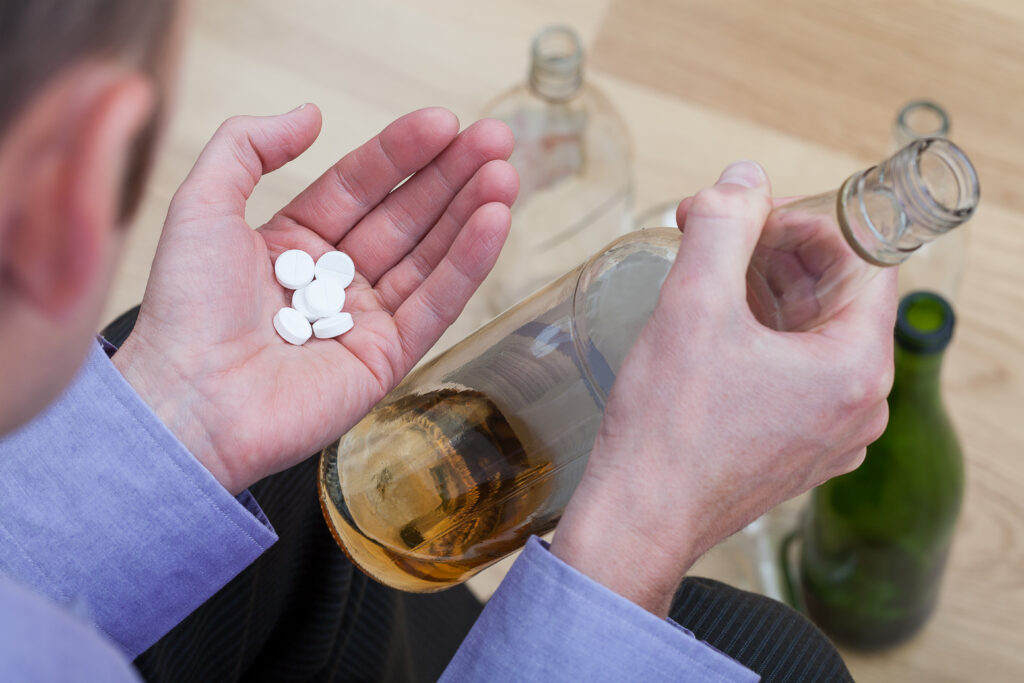A man holding a bottle of alcohol in one hand and Adderall pills in the other as he contemplates the relationship between alcohol and Adderall.