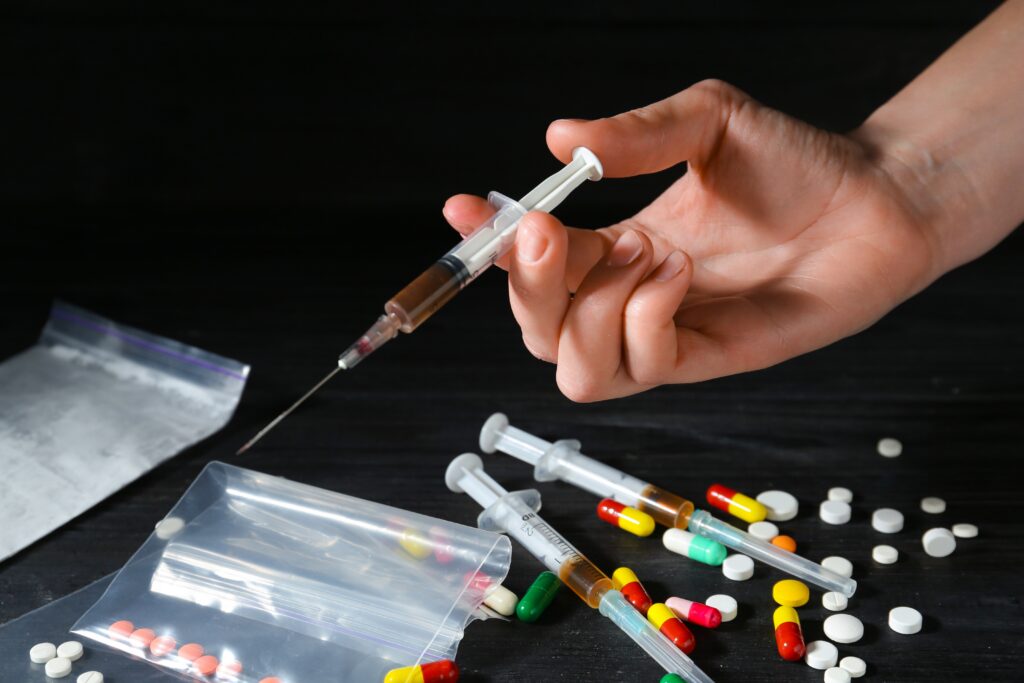 A person holding a syringe of drugs surrounded by bags and pills to fuel their amphetamine addiction.